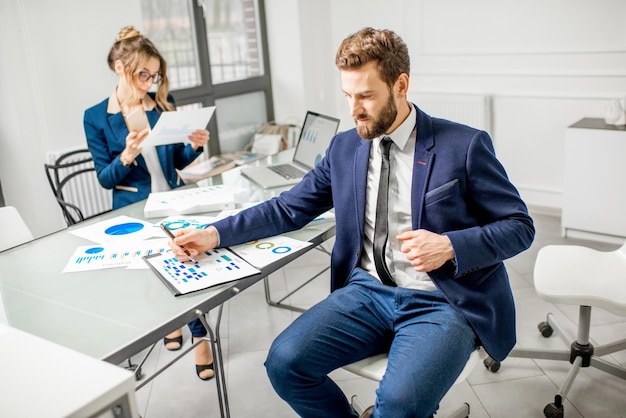 Ritratto di un uomo d'affari vestito in abito che lavora su documenti con assistente femminile sullo sfondo all'interno dell'ufficio bianco