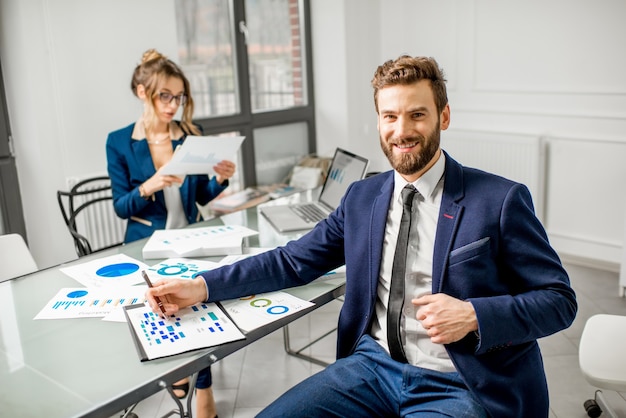 Ritratto di un uomo d'affari vestito in abito che lavora su documenti con assistente femminile sullo sfondo all'interno dell'ufficio bianco