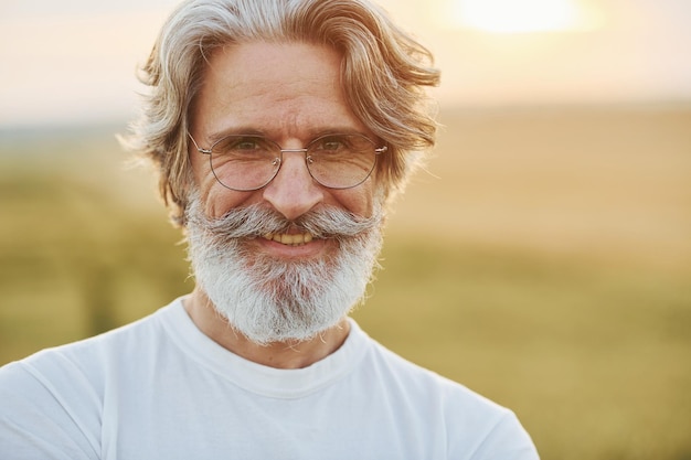 Ritratto di un uomo elegante con capelli grigi e barba che si trova all'aperto sul campo in una giornata di sole