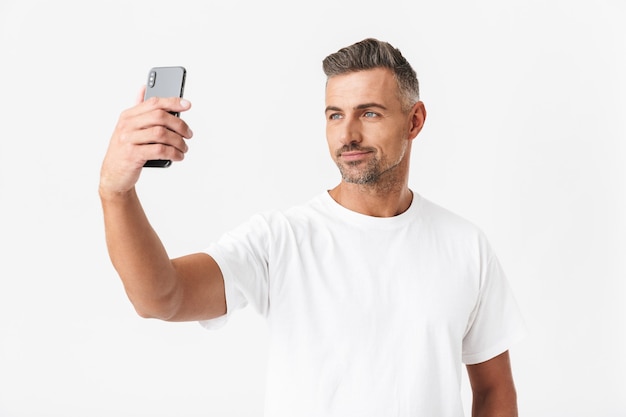 Ritratto di un uomo felice di 30 anni che indossa una maglietta casual che si fa selfie isolato su bianco