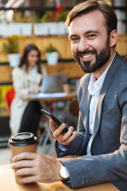 Ritratto di un uomo felice sorridente che indossa una giacca che beve caffè da asporto e usa il telefono cellulare mentre è seduto al bar