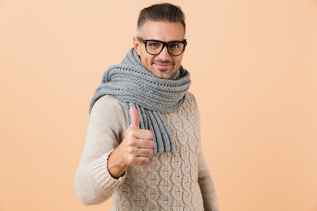 Ritratto di un uomo fiducioso vestito in maglione e sciarpa in piedi isolato sopra il muro beige, dando i pollici in su