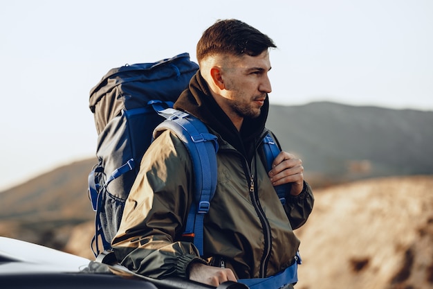 Ritratto di un uomo giovane viaggiatore in attrezzatura da escursionismo in piedi vicino alla sua auto fuoristrada all'alba