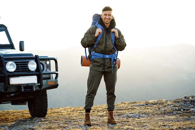 Ritratto di un uomo giovane viaggiatore in attrezzatura da trekking in piedi vicino alla sua auto fuoristrada all'alba