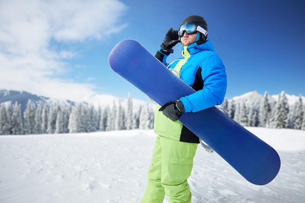 Ritratto di un uomo in una stazione sciistica sullo sfondo montagne che tiene una tavola da snowboard indossando occhiali da sci