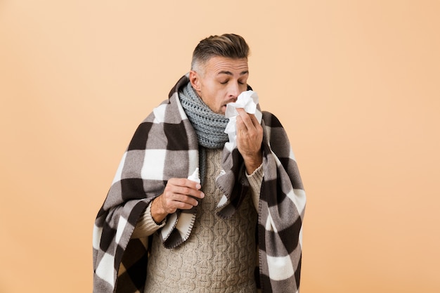 Ritratto di un uomo malato avvolto in una coperta in piedi isolato sopra il muro beige, avendo il naso che cola, usando il tovagliolo