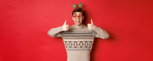 Ritratto di un uomo soddisfatto e felice con un maglione natalizio e occhiali da festa che mostra pollice in su che desidera ...