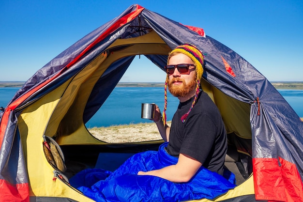 Ritratto di un uomo turistico elegante con barba rossa e zenzero in un cappello colorato fatto di lana di yak dal Nepal che tiene una tazza di tè e caffè in un sacco a pelo avvolto in tenda sullo sfondo del lago