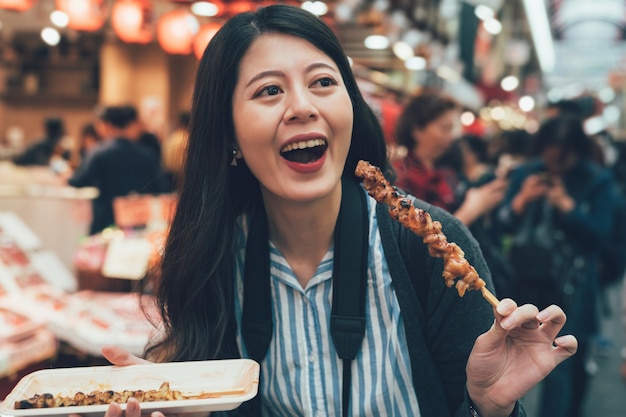 Ritratto di un visitatore asiatico felice che visita il mercato locale giapponese di kuromon ichiba a osaka in giappone mangiando deliziosi yakitori sulla strada. giovane donna sorridente dopo aver provato gustoso pollo al barbecue.