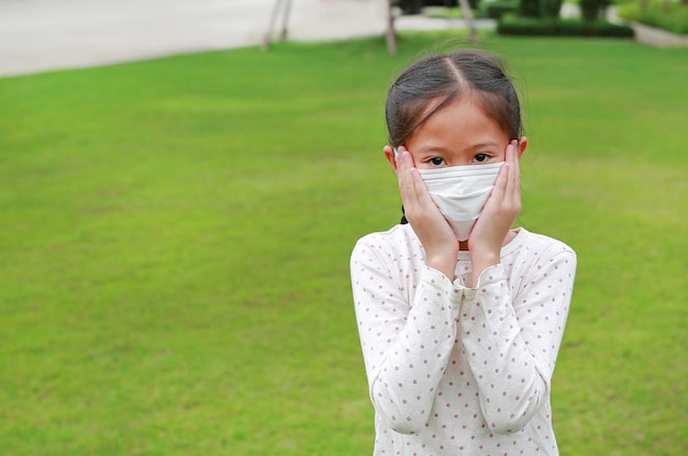 Ritratto di una bambina asiatica che indossa una maschera medica e tocca le guance per proteggere il covid19 mentre resta in giardino