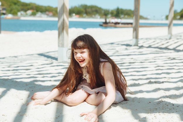 Ritratto di una bambina bionda in un vestito seduto sulla sabbia della spiaggia Giornata di sole estivo