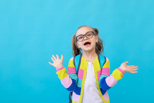 Ritratto di una bambina carina con gli occhiali in una giacca a righe con quaderni in mano e uno zaino che si rallegra e si diverte. Il concetto di educazione. sfondo blu, posto per il testo