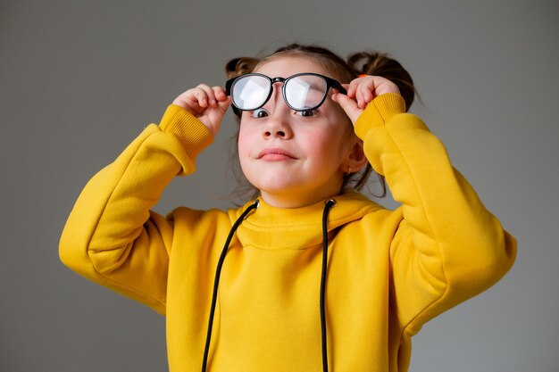 Ritratto di una bambina carina e divertente con occhiali con montatura nera in felpa con cappuccio gialla con un'acconciatura divertente