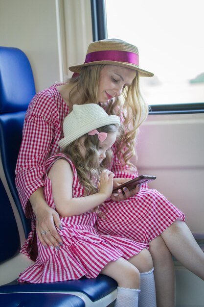 Ritratto di una bambina caucasica con sua mamma in cappelli con capelli ondulati che usa uno smartphone in treno