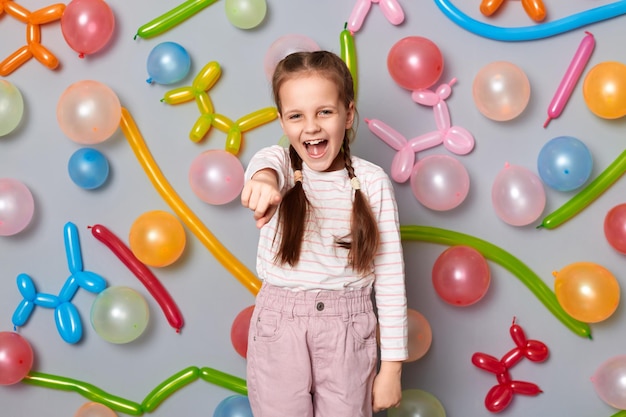 Ritratto di una bambina che ride con le trecce che indossa abiti casual in piedi contro il muro grigio con palloncini colorati che ti prendono in giro puntando il dito indice verso la fotocamera