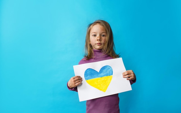 Ritratto di una bambina con la faccia sporca della guerra in piedi su uno sfondo blu con in mano un cuore dipinto con i colori della bandiera dell'Ucraina