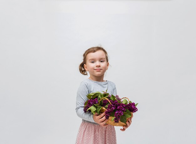 Ritratto di una bambina con un cesto di lillà. Ragazza carina in un abito grigio con una coda su uno spazio bianco. Copia spazio