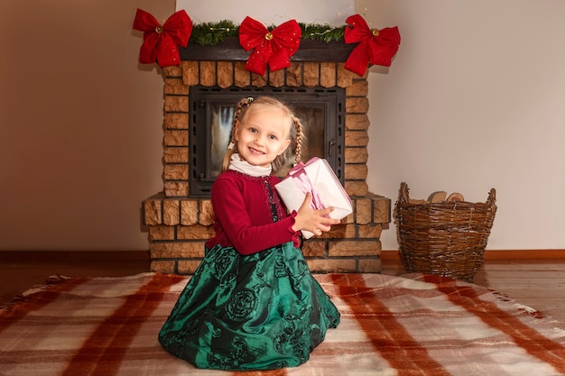 Ritratto di una bambina con un vestito verde e un maglione rosso con un regalo in mano sullo sfondo di Natale