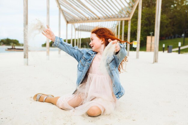 Ritratto di una bambina dai capelli rossi in un vestito seduto sulla sabbia della spiaggia Giornata di sole estivo