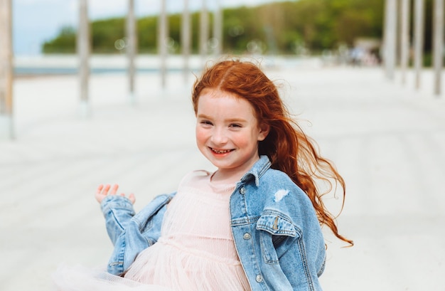 Ritratto di una bambina dai capelli rossi in un vestito seduto sulla sabbia della spiaggia Giornata di sole estivo