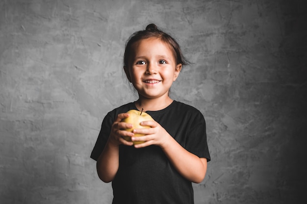 Ritratto di una bambina di felicità che mangia una mela verde sul muro grigio