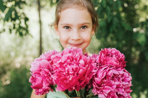 Ritratto di una bambina di sette anni caucasica carina felice, tiene in mano un mazzo di fiori di peonia rosa in piena fioritura sullo sfondo verde della natura