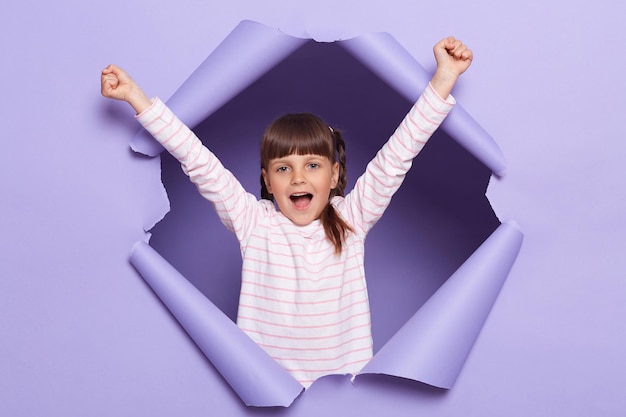 Ritratto di una bambina eccitata stupita con trecce che indossano una camicia a righe in posa in una parete di carta strappata bambino in piedi con le braccia alzate che urlano felicemente celebrando il suo successo