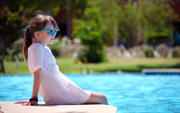 Ritratto di una bambina felice che si rilassa sul lato della piscina nelle soleggiate giornate estive durante le vacanze tropicali