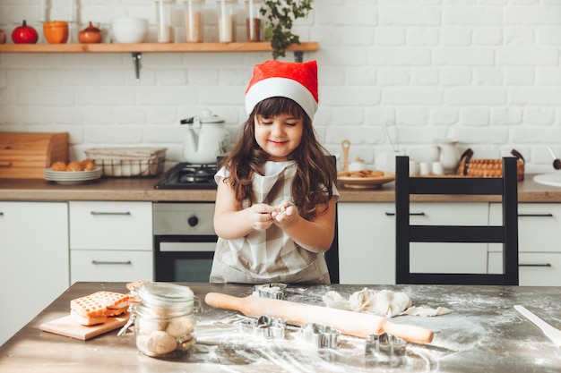 Ritratto di una bambina felice con un cappello da Babbo Natale che stende la pasta sul tavolo della cucina un bambino che prepara i biscotti di Natale