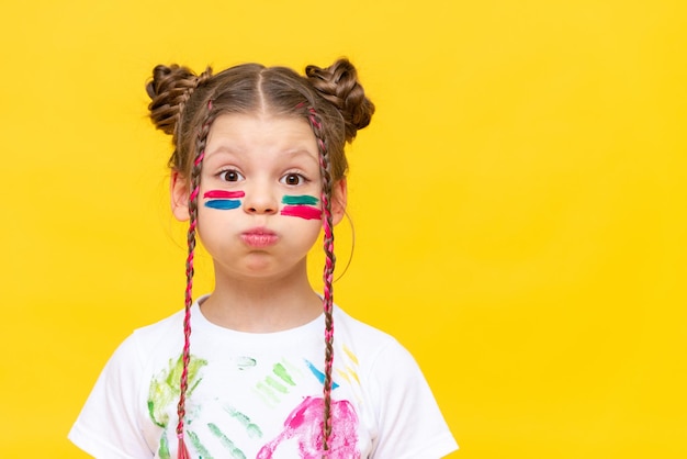 Ritratto di una bambina imbrattata di vernici multicolori per disegnare su uno sfondo giallo isolato La studentessa gonfiò le guance Sviluppo della creatività e dell'arte dei bambini