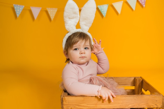 Ritratto di una bambina in un abito rosa con orecchie di coniglio seduto in una scatola di legno su uno sfondo giallo