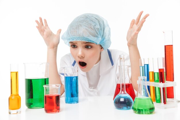 Ritratto di una bambina perplessa in camice bianco da laboratorio che fa esperimenti chimici con liquido multicolore in provette isolate su un muro bianco