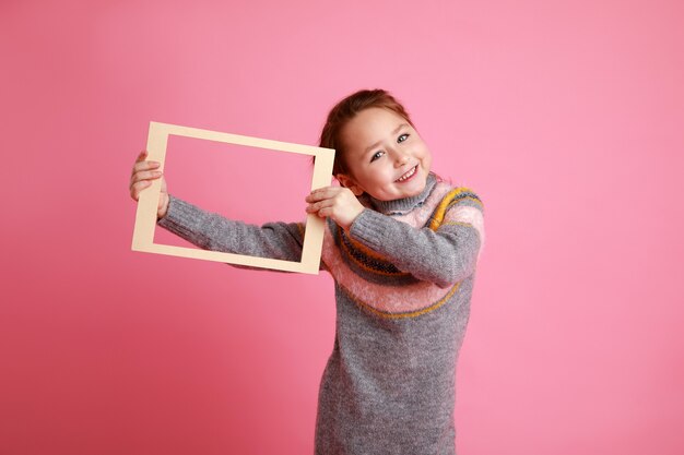 Ritratto di una bambina sorridente che tiene una cornice vuota per un mock-up su uno sfondo rosa.