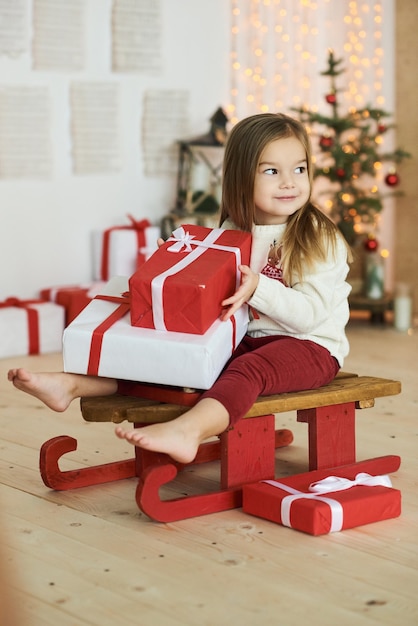 Ritratto di una bambina su una slitta rossa con un regalo su uno sfondo di luci sfocate