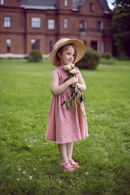 Ritratto di una bella bambina in un vestito rosa e cappello in piedi con un mazzo di fiori pioni su un prato verde in estate