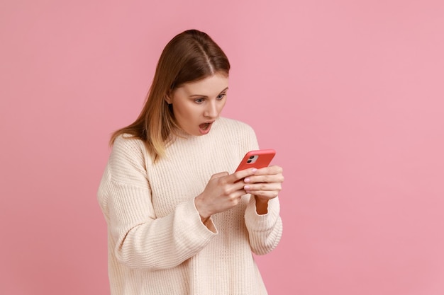 Ritratto di una bella blogger bionda stupita che usa lo smartphone, guardando lo schermo del dispositivo, vede contenuti scioccanti, indossa un maglione bianco. Studio indoor girato isolato su sfondo rosa.