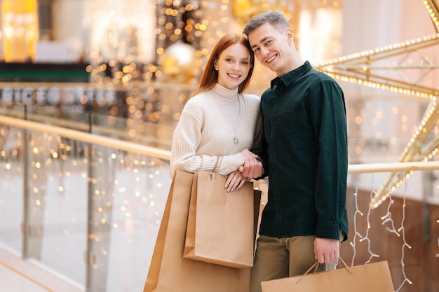 Ritratto di una bella coppia giovane e allegra che tiene in mano le borse della spesa abbracciate in piedi nel centro commerciale sfocato