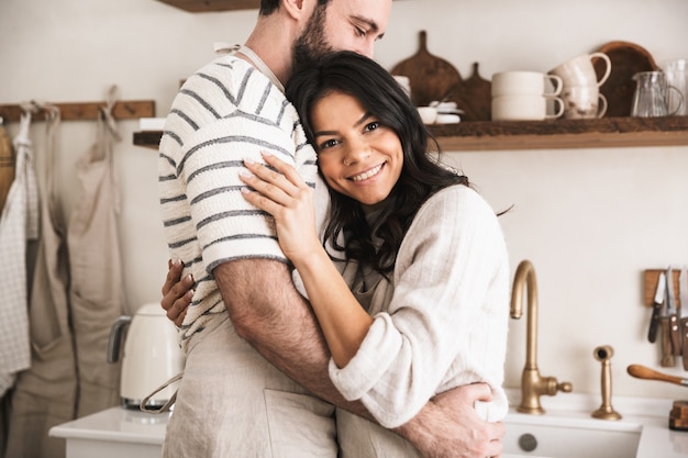 Ritratto di una bella coppia uomo e donna di 30 anni che indossa grembiuli che si abbracciano mentre cucinano in cucina a casa