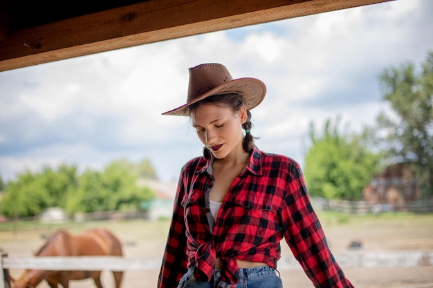 Ritratto di una bella cowgirl vestita con una camicia rossa e un cappello sullo sfondo di una stalla