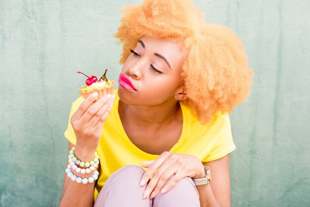 Ritratto di una bella donna africana in maglietta gialla che tiene un dolce dessert con ciliegia sullo sfondo verde