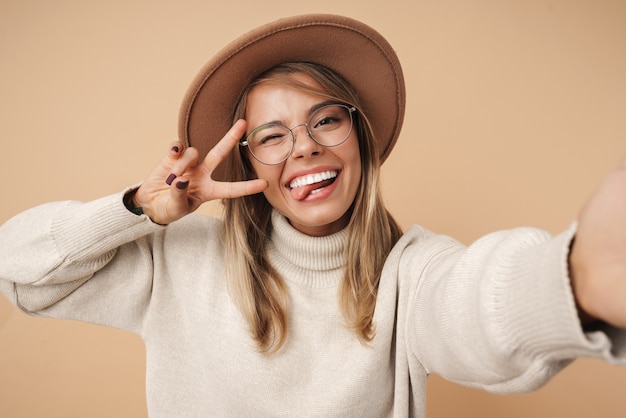 Ritratto di una bella donna allegra con un cappello che fa un gesto di pace e scatta una foto selfie