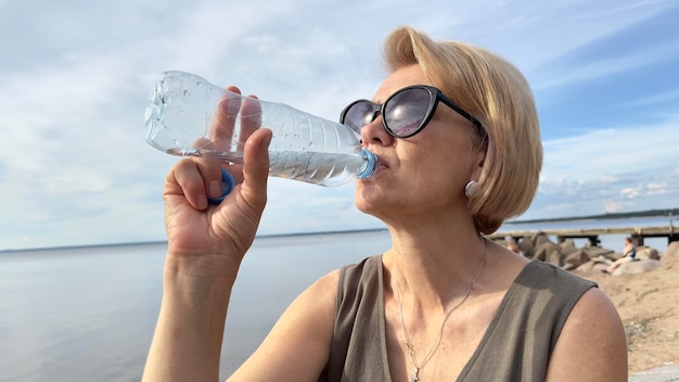 Ritratto di una bella donna anziana che beve acqua da una bottiglia di plastica durante il giorno estivo