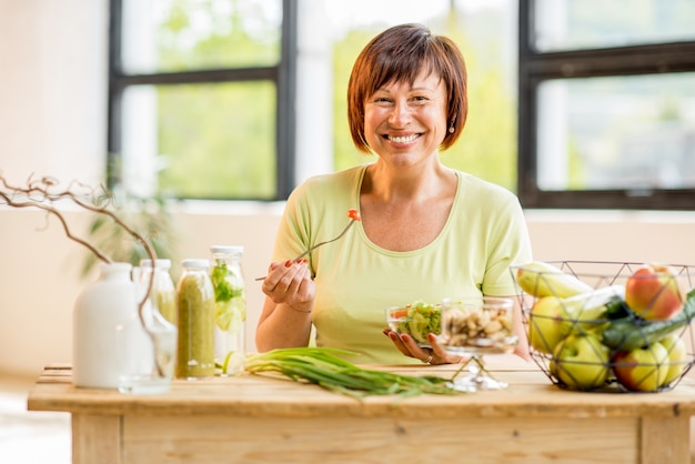 Ritratto di una bella donna anziana con cibo sano verde sul tavolo al chiuso sullo sfondo della finestra