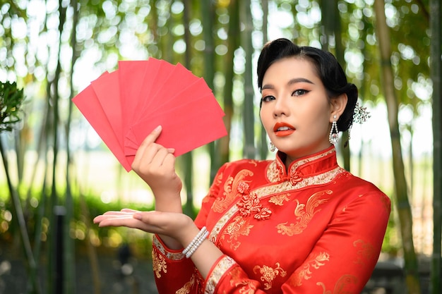 Ritratto di una bella donna asiatica in un cheongsam cinese in posa con buste rosse uomo e palme aperte nella foresta di bambù