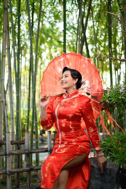 Ritratto di una bella donna asiatica in un cheongsam cinese in posa con un bellissimo ombrello di carta rosso sulla foresta di bambù, copia spazio