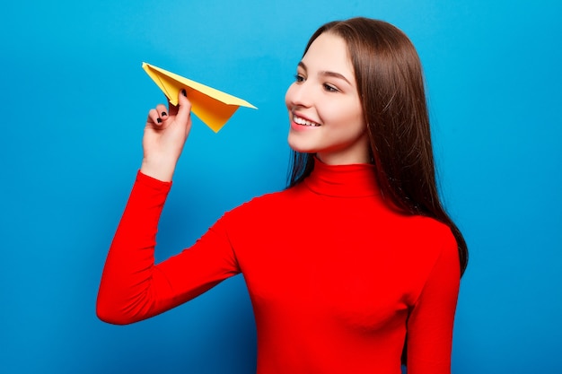 Ritratto di una bella donna che tiene un aeroplano di carta giallo. Su sfondo blu.