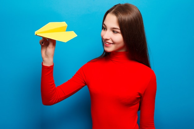 Ritratto di una bella donna che tiene un aeroplano di carta giallo. Su sfondo blu.