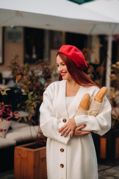 Ritratto di una bella donna con un berretto rosso e un cardigan bianco con baguette in mano passeggiando per la città autunnale