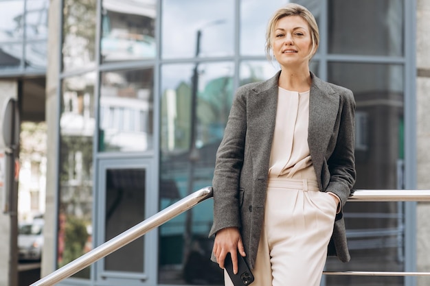 Ritratto di una bella donna d'affari matura in giacca e cravatta grigia che sorride e parla al telefono sullo sfondo degli edifici urbani e per uffici moderni