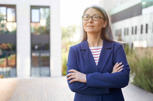 Ritratto di una bella donna d'affari matura sicura di sé in occhiali e abbigliamento classico che tiene le braccia incrociate e guarda da parte mentre si trova contro l'edificio per uffici all'aperto. Concetto di affari